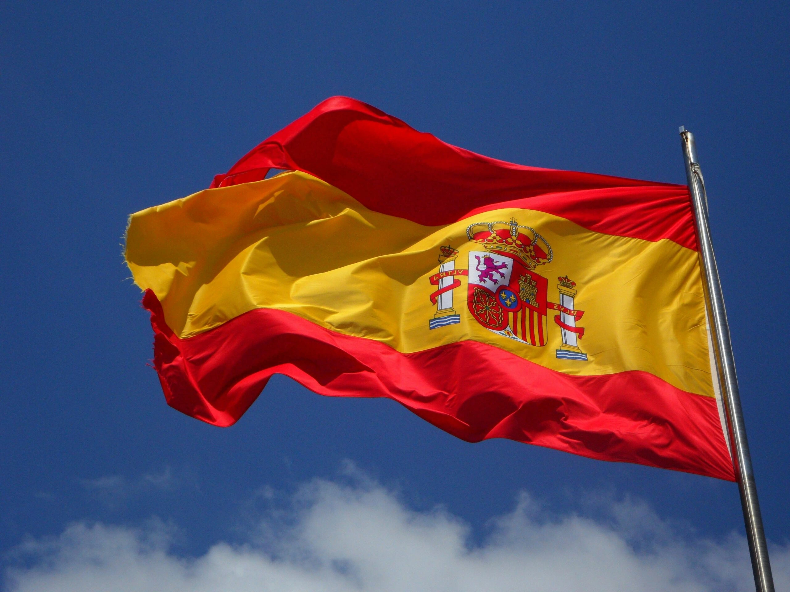 The national flag of Spain is waving vigorously on a flagpole against a bright blue sky with a few white clouds