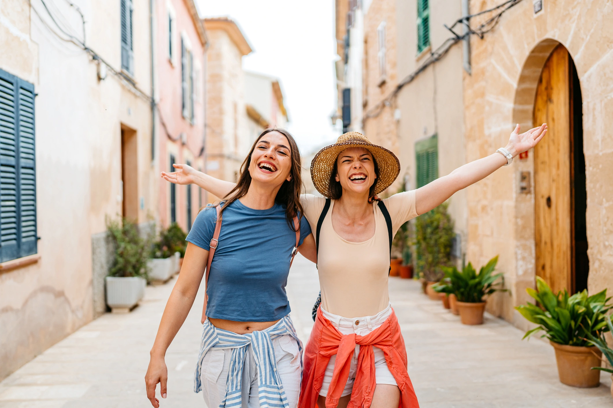 Two women are smiling and walking with their arms outstretched down a narrow, historic street.