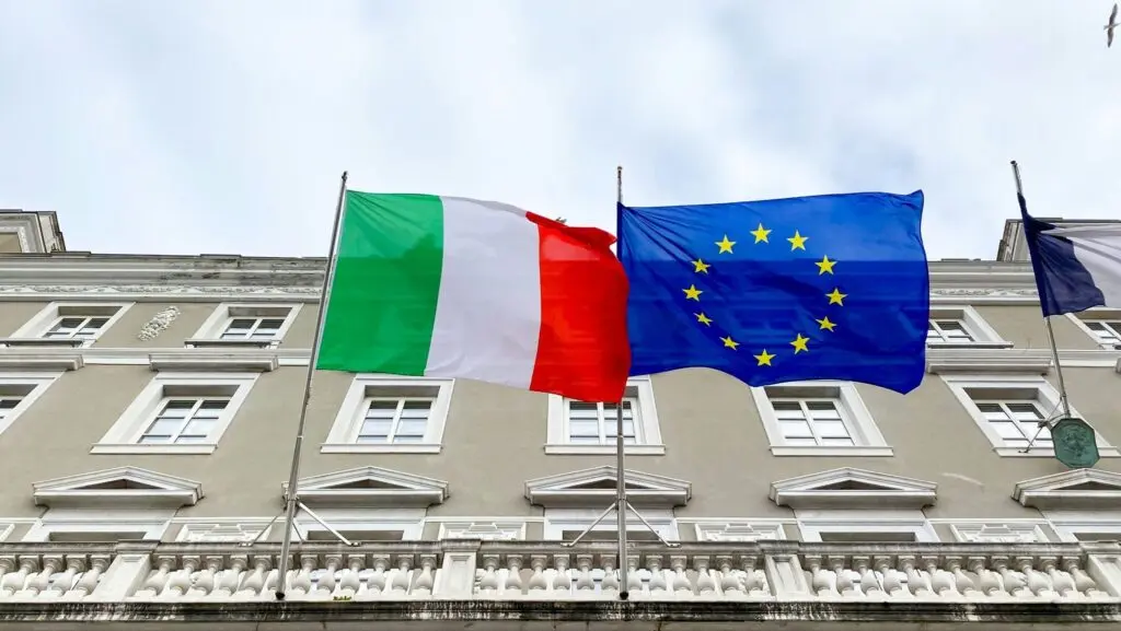 An Italian flag and an EU flag fly against a building backdrop; Italy's Supreme Court is set to rule on citizenship reform retroactivity in January 2026, affecting Italian citizenship by descent cases.
